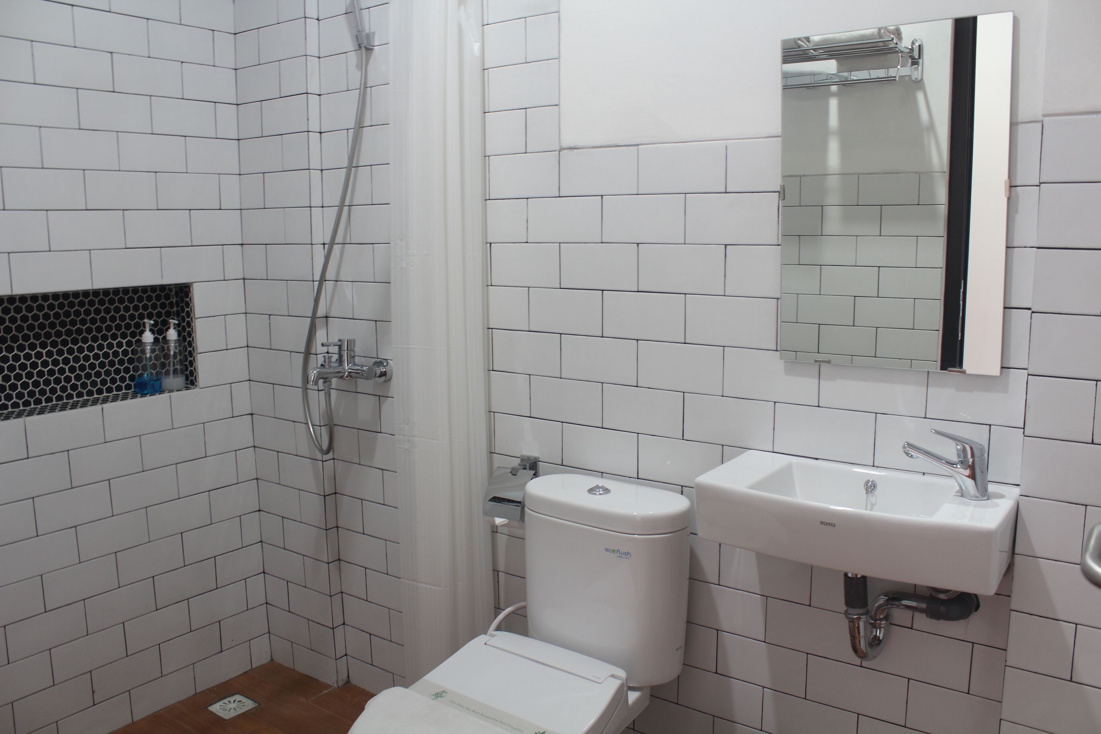 A bathroom with white tiled walls, a white toilet, a sink with a mirror above it, and a shower area.