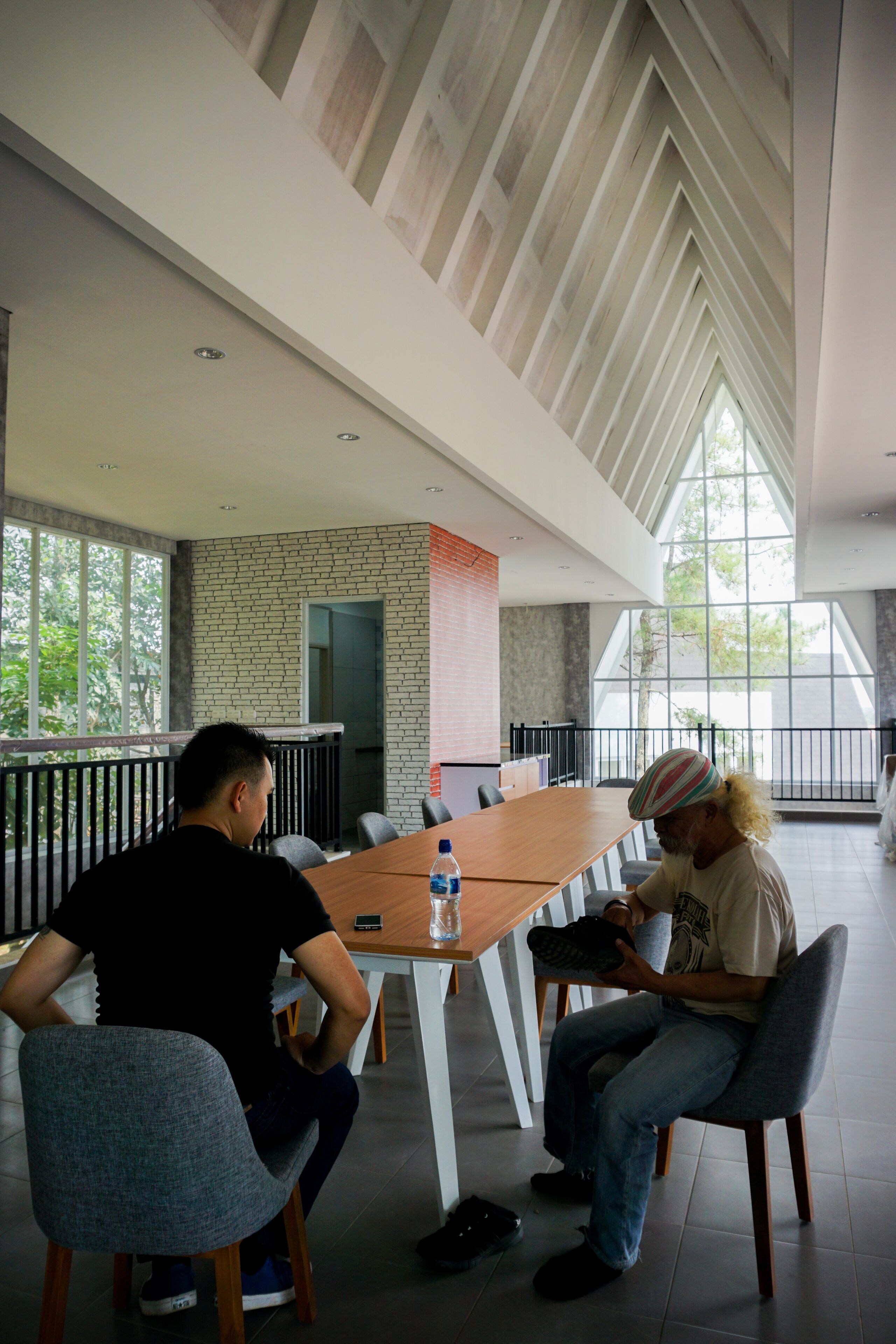 A modern indoor space with a high ceiling, large windows, and a long table with chairs.