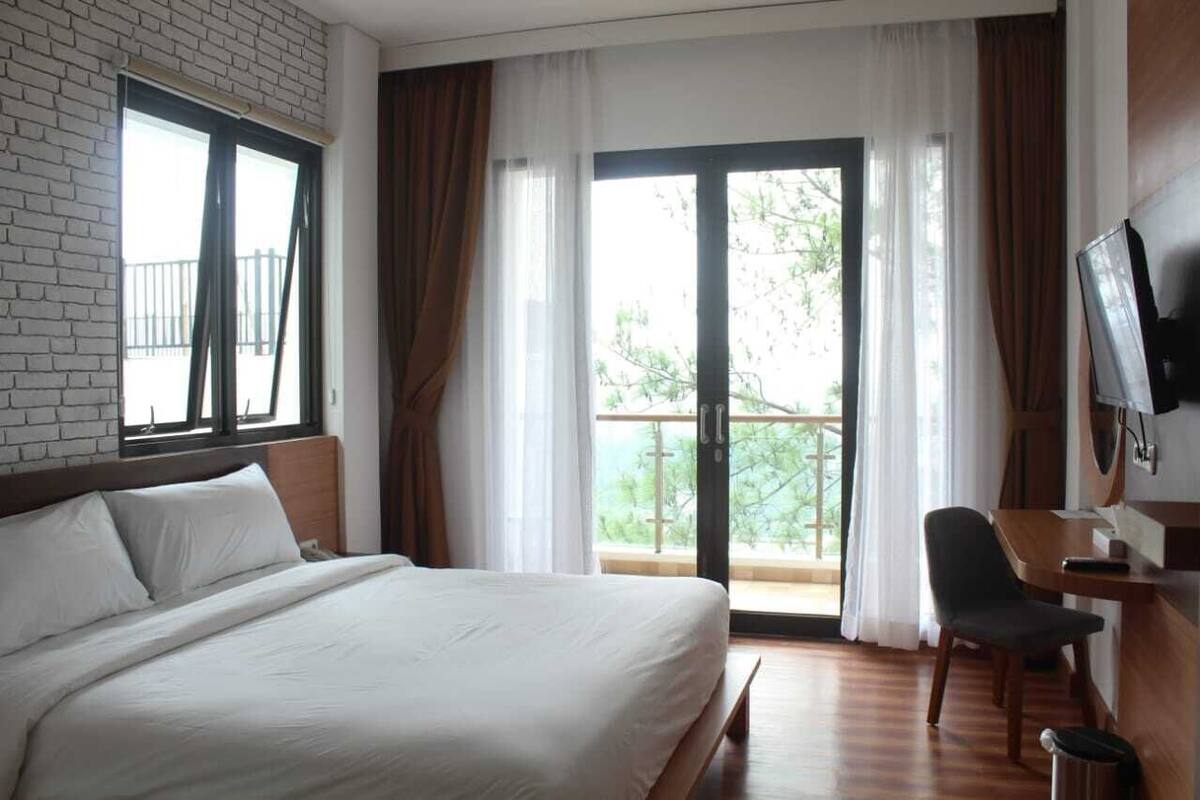 A hotel room with a large bed, a desk, a chair, a TV, and a balcony with curtains.