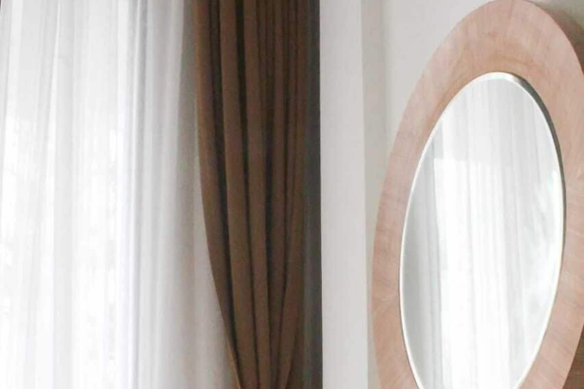 A modern room with a grey upholstered chair, a wooden vanity with a round mirror, and a window with sheer curtains.