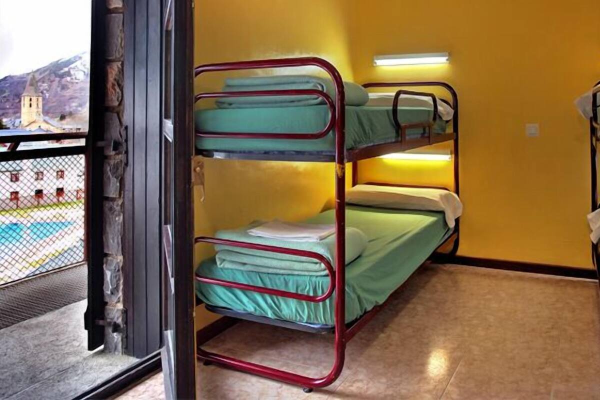 A bunk room with three bunk beds, a window with a view of a building and mountains, and a door leading outside.