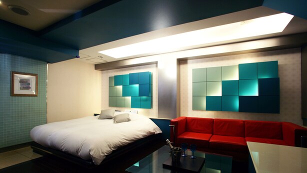 A modern hotel room with a bed, a red sofa, a glass coffee table, and a wall with a geometric light fixture. - Hotel AQUA Blue Yokosuka - Adults Only (Yokosuka)