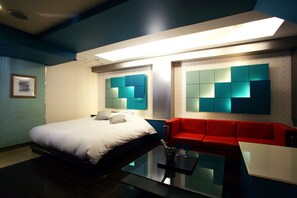 Une chambre d’hôtel moderne avec un lit, un canapé rouge, une table basse en verre et un mur agrémenté d’un luminaire aux motifs géométriques.