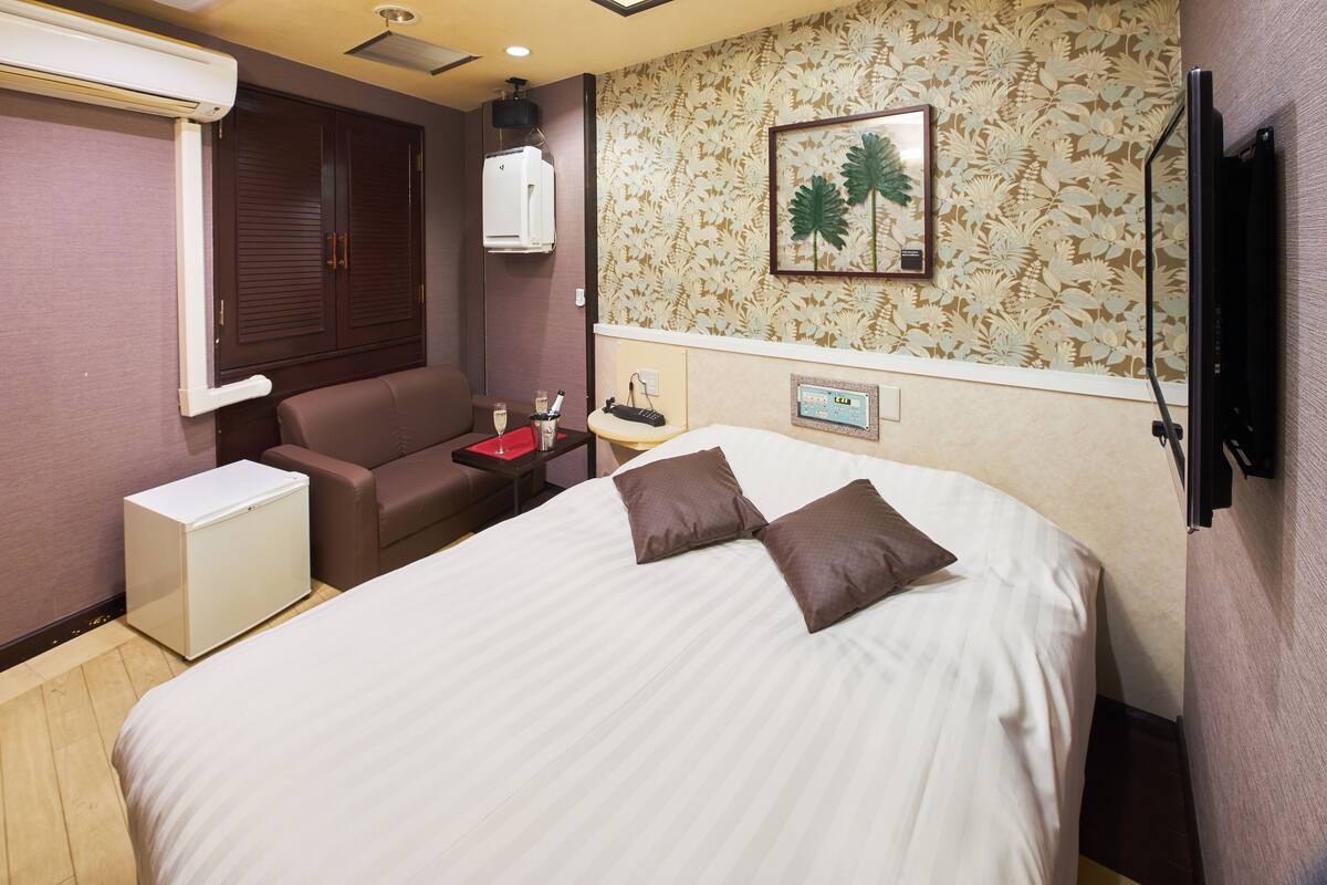 Ein Hotelzimmer mit Bett, kleinem Schreibtisch, Stuhl, Kleiderschrank und einem an der Wand befestigten Fernseher.