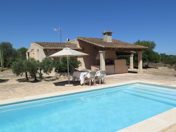 Un espace aménagé au bord de la piscine, comprenant un coin salon couvert, un barbecue et une maison avec un toit en tuiles.