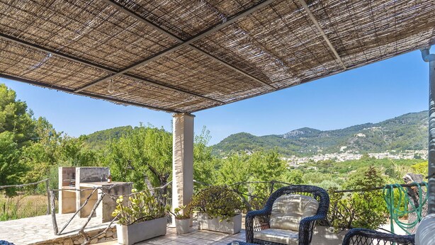 A covered outdoor seating area with wicker furniture, potted plants, and a view of a hillside town.