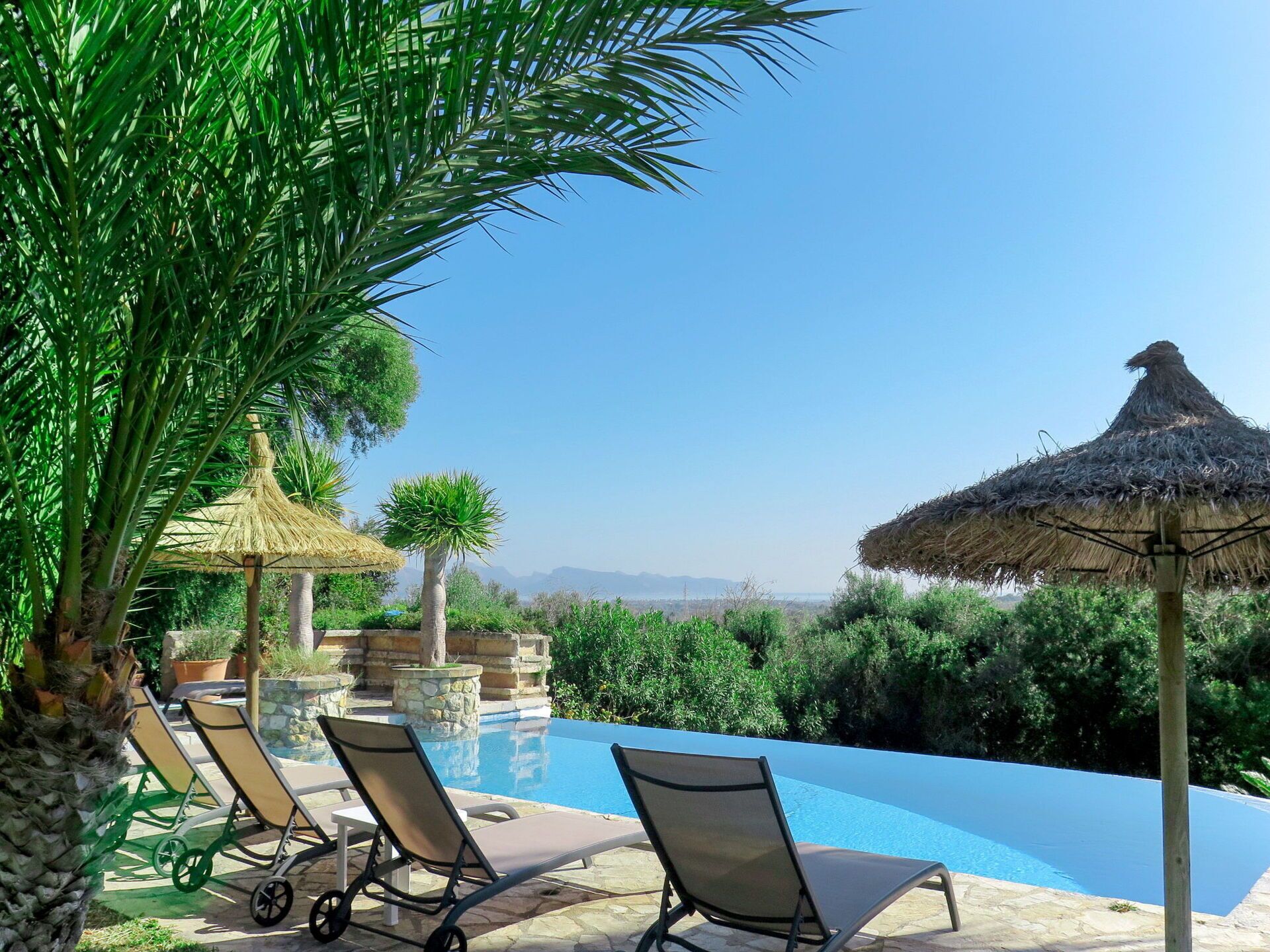 Ein Poolbereich mit Liegestühlen und einem Reetdach-Sonnenschirm, umgeben von Grünflächen und blauem Himmel.