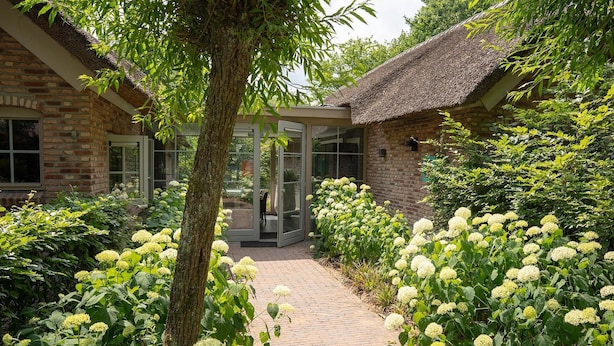 A brick building with a glass door entrance, surrounded by lush greenery and white flowers. - Vlindervallei Studio 2p (Ermelo)
