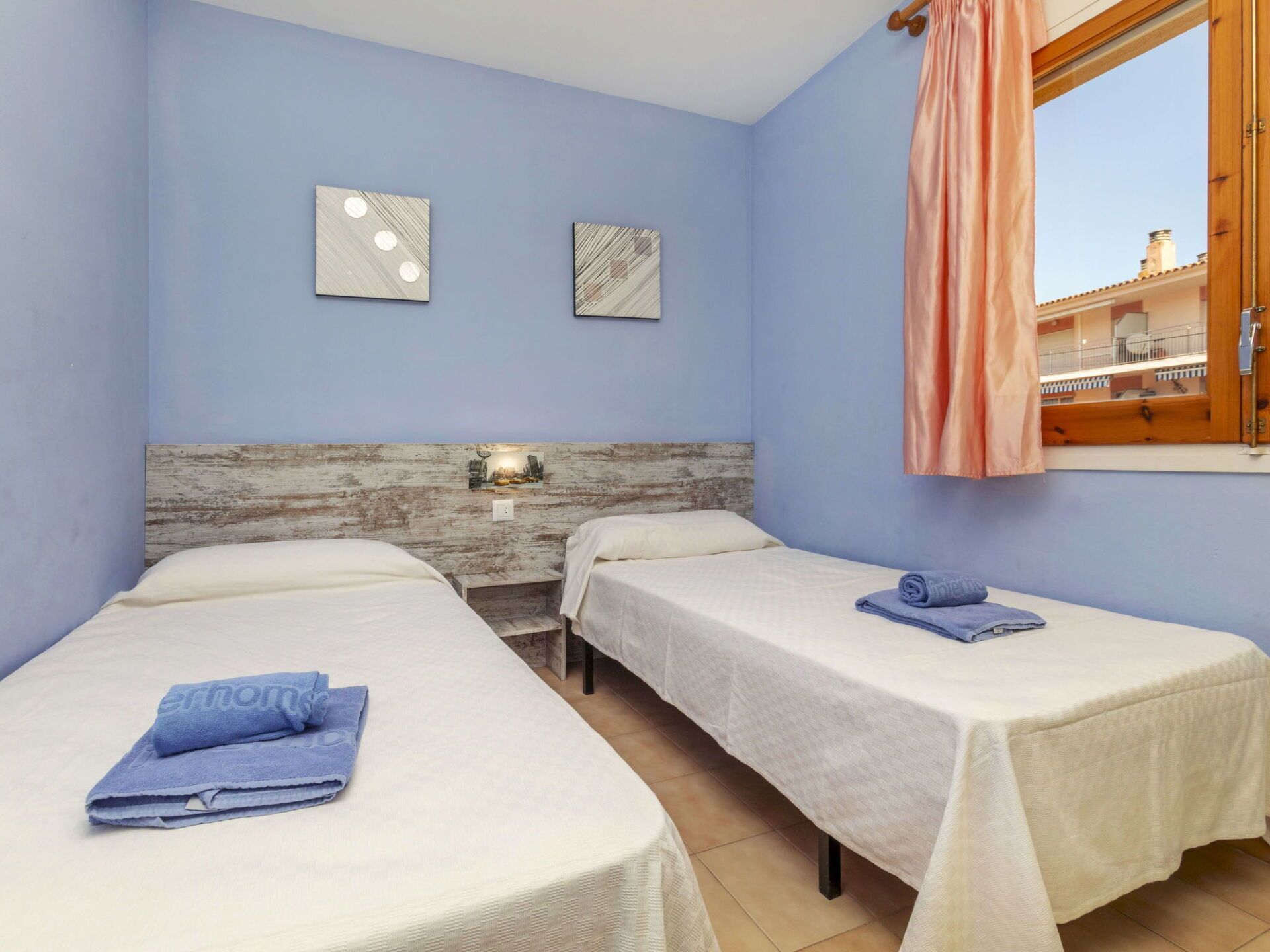 Une chambre avec deux lits, une tête de lit en bois, une fenêtre avec un rideau et deux appliques murales.