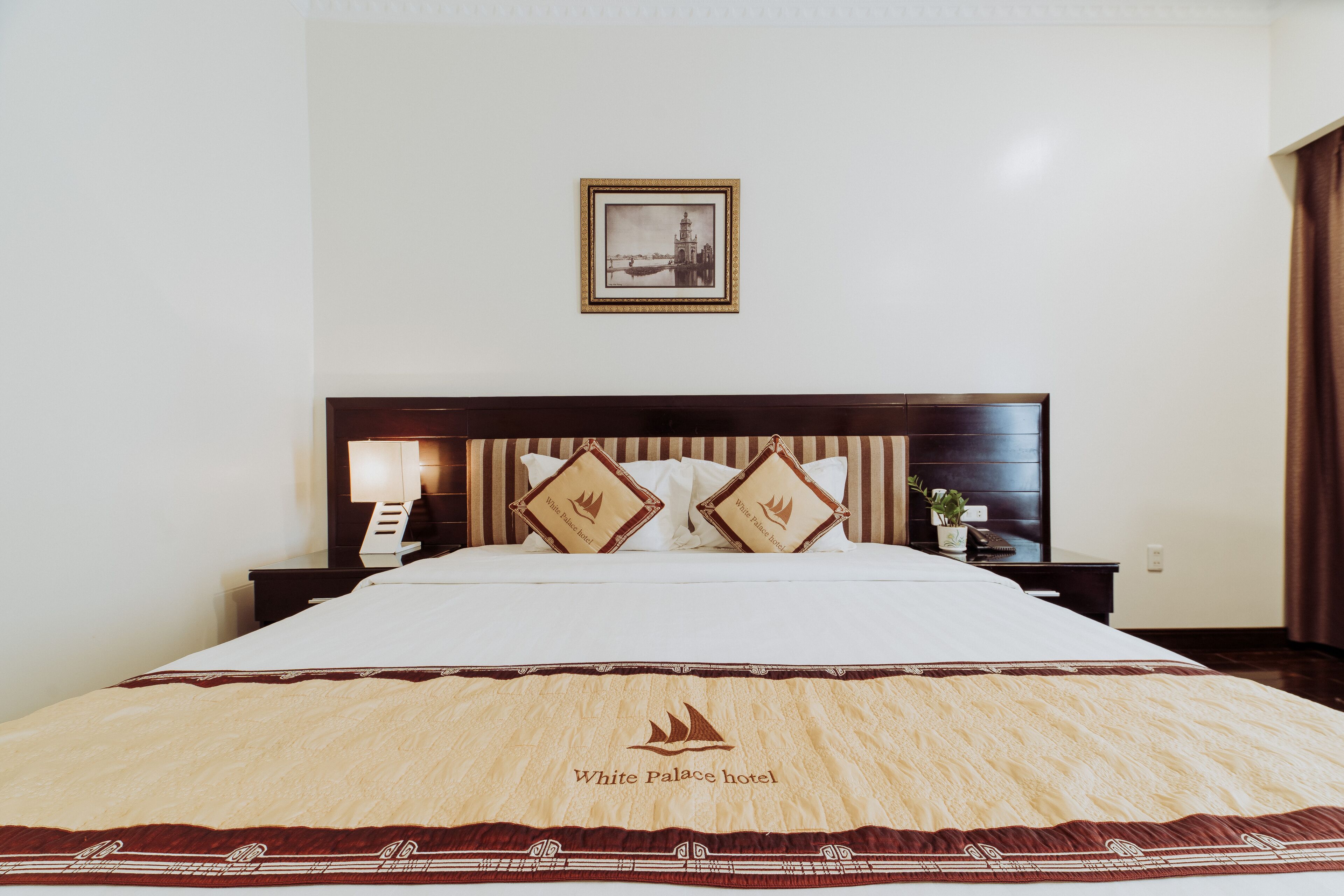 A hotel room with a large bed, a headboard with a framed picture, two decorative pillows, a bedside table with a lamp, and a wall-mounted picture.