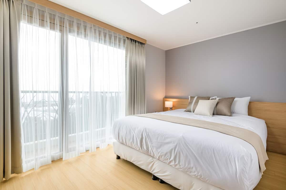 A modern hotel room with a large bed, a nightstand with a lamp, sheer curtains, and a view of a balcony.