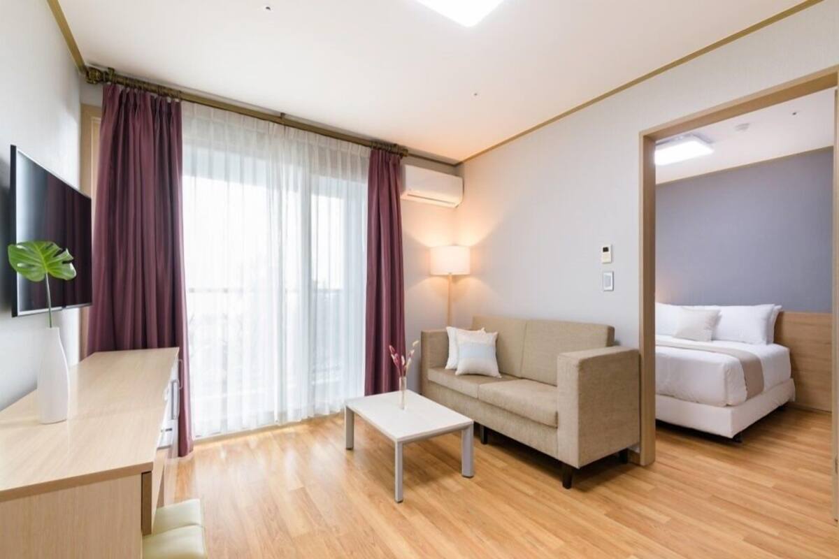 A modern hotel room with a wooden floor, a beige sofa, a small white table, a TV on the left, and a bed with white bedding.