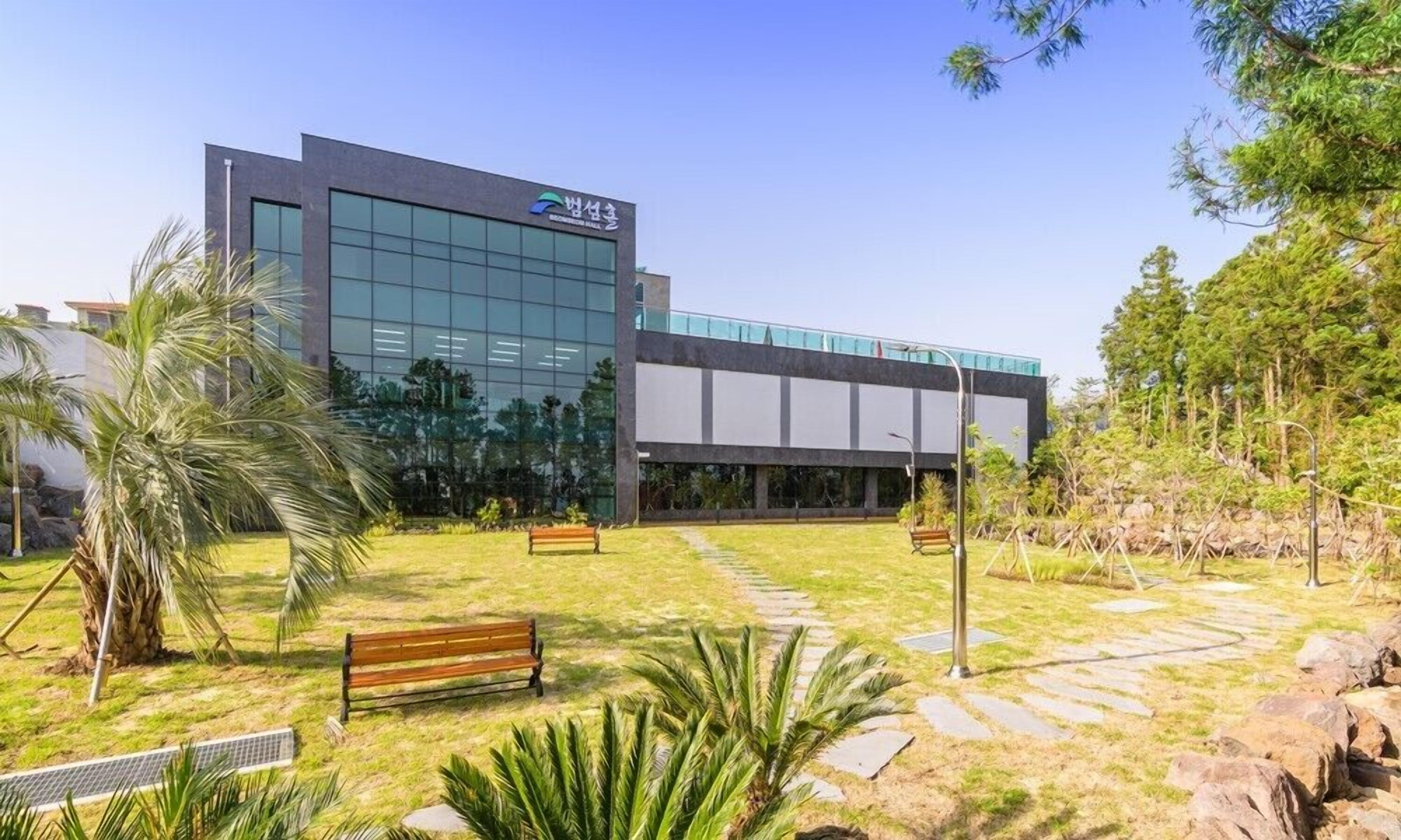 A modern building with a glass facade, a paved courtyard with a bench, and tropical landscaping.