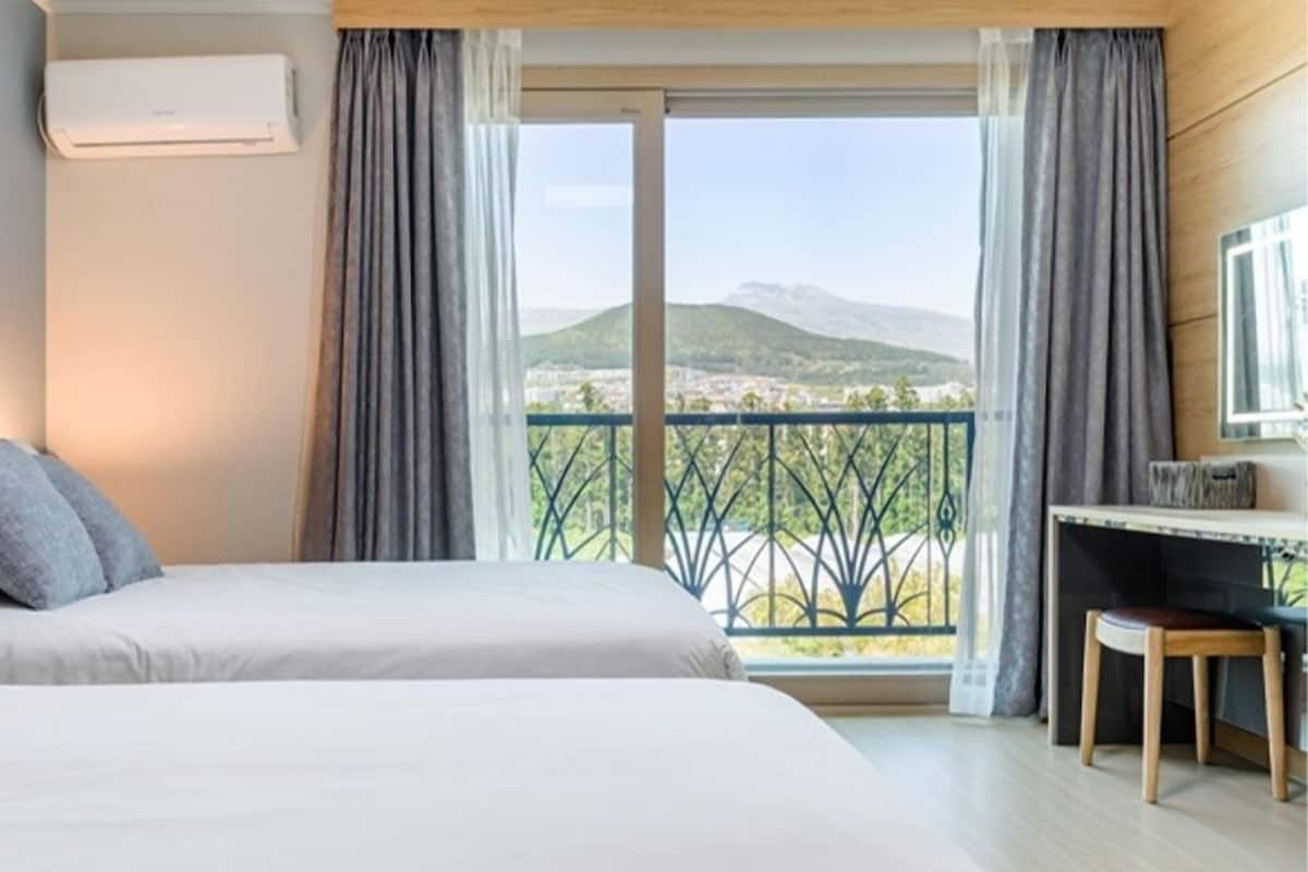 A hotel room with two beds, a desk, and a view of a balcony with a railing.
