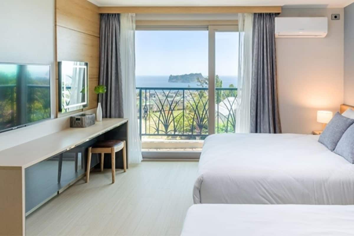 A modern hotel room with a large bed, a desk with a chair, a TV, and a balcony with a view of the sea.