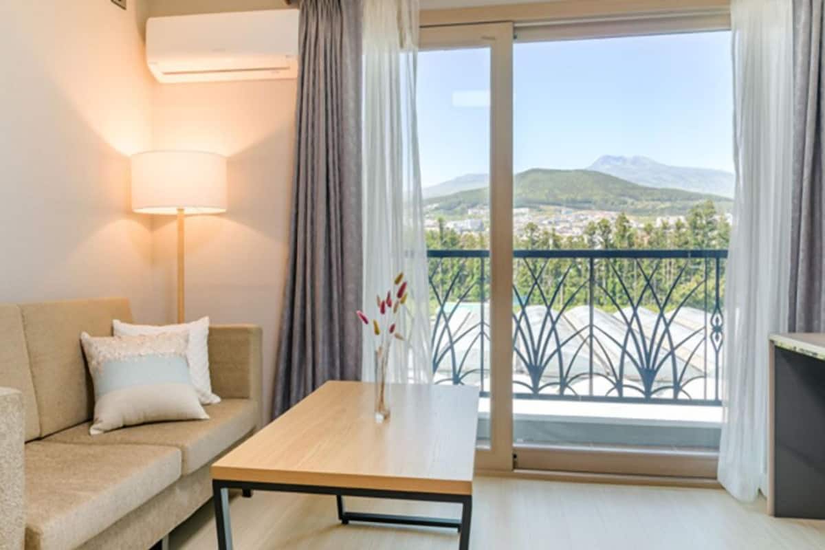 A modern hotel room with a sofa, a table, and a view of a cityscape through a balcony.