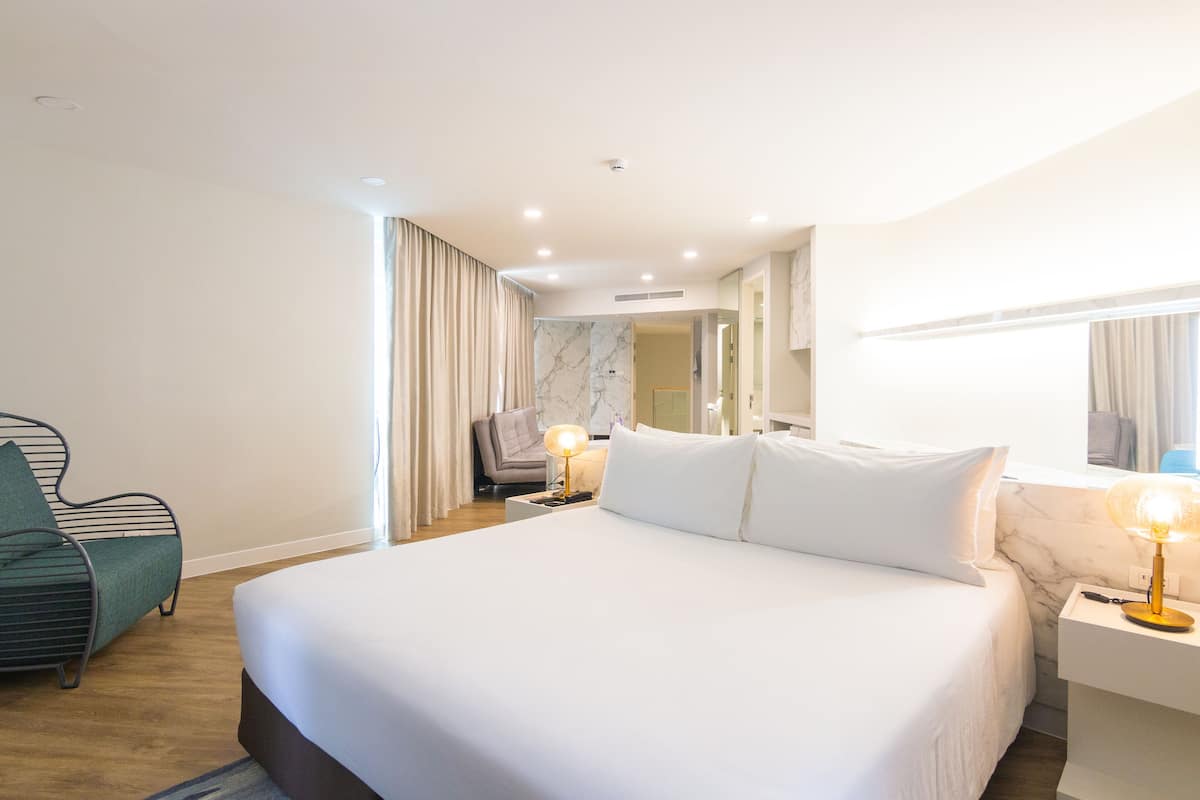 Habitación de hotel con una cama grande, una mesita de noche con lámpara, una silla y un baño visible a través de una puerta de cristal.