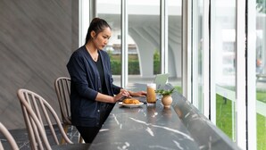 Henkilö työskentelee pöydän ääressä, ja pöydällä on kannettava tietokone, lautanen ruokaa ja juoma. Taustalla on moderni ympäristö, jossa on suuret ikkunat.