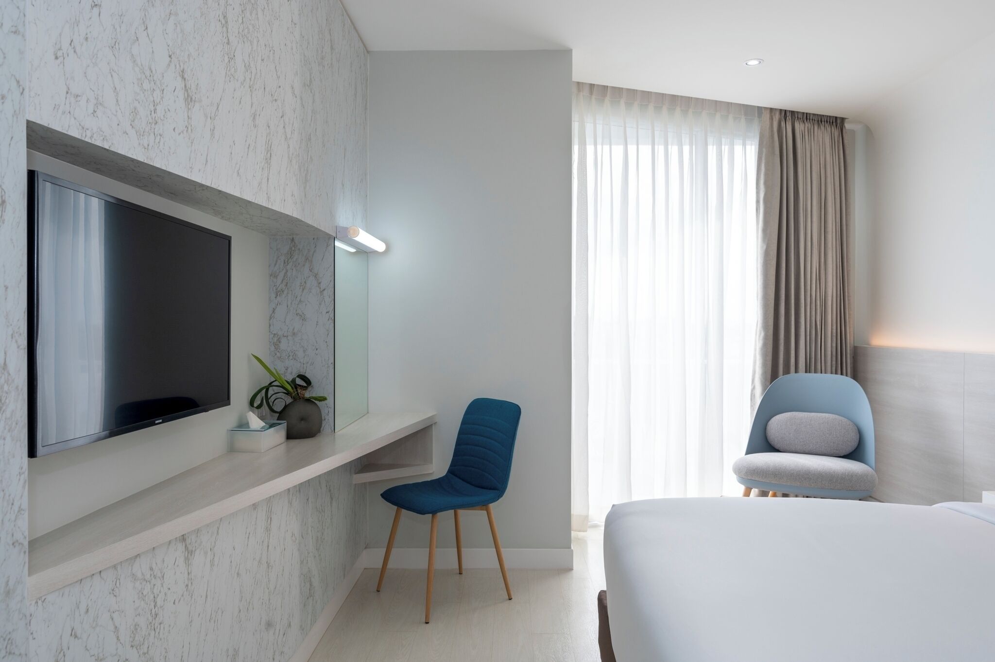 Habitación de hotel moderna con un televisor de pantalla plana grande montado en una pared de mármol, una silla azul, una cama blanca y un ventanal con cortinas.