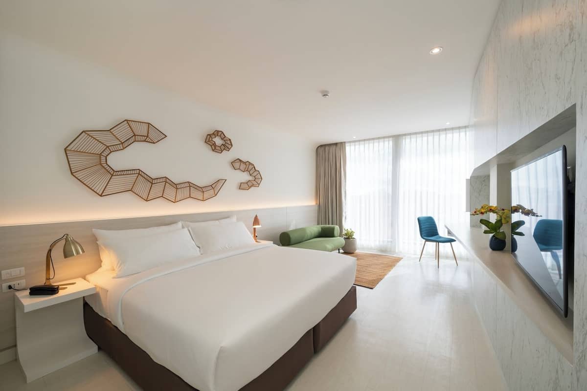 Una habitación de hotel moderna con una cama grande, una mesita de noche con lámpara, una silla verde y un ventanal con cortinas.