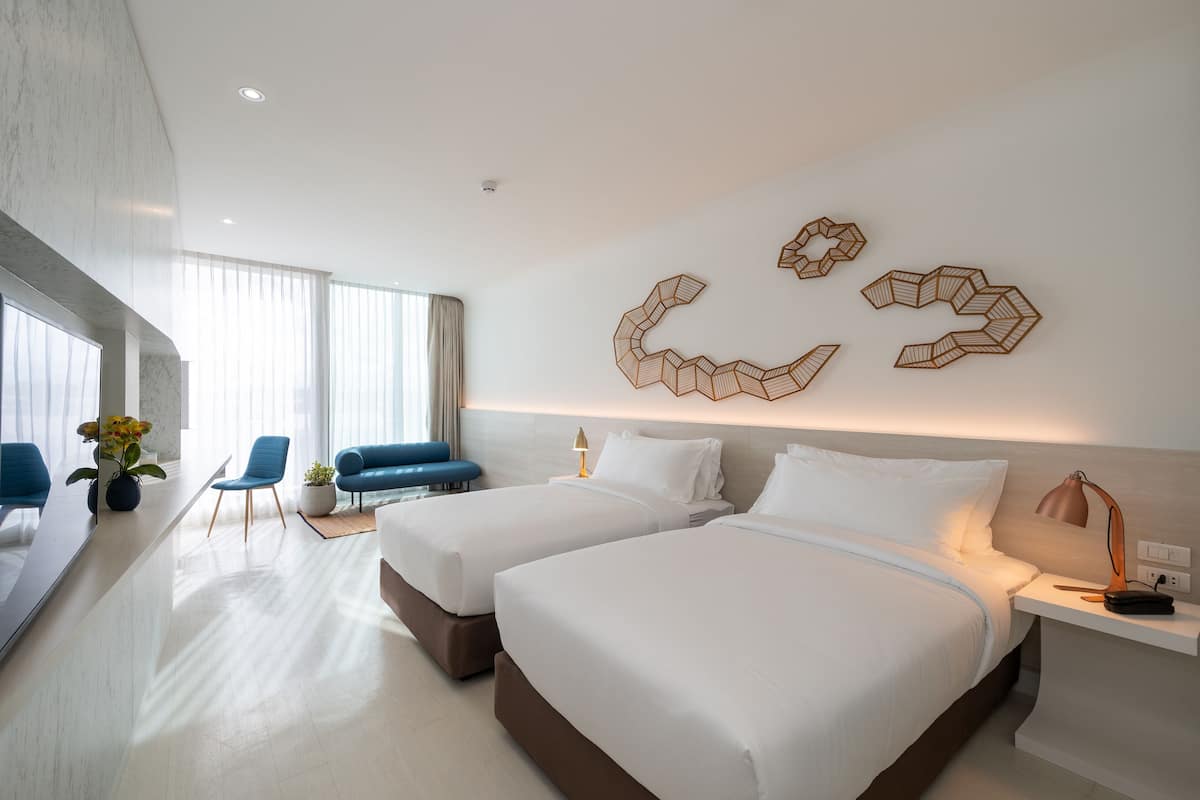 Una habitación de hotel moderna con dos camas, una zona de estar con sillas, un espejo grande y decoración mural.