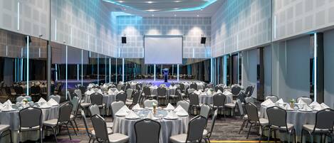 A large, well-lit conference room with round tables and chairs, a stage, and a large screen.