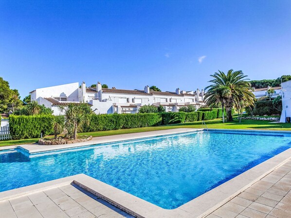 Une piscine à l’eau d’un bleu limpide, entourée d’une terrasse dallée. Au fond, on aperçoit des bâtiments blancs aux toits recouverts de tuiles rouges et une pelouse bien entretenue agrémentée de palmiers.