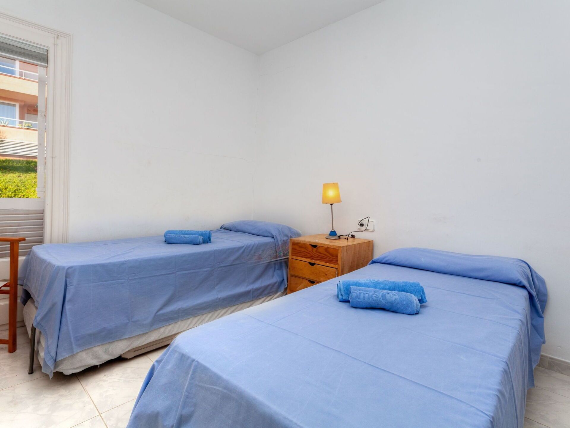 A small, clean room with two single beds, a wooden nightstand with a lamp, and a window with blinds.