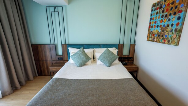 A neatly made bed with pillows, a bedside table, a wall art piece, and a window with curtains. - Leaf Port Hotel (Konyaalti)