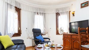 A living room with a black leather sofa, a wooden dining table with cups and a cake, a black armchair with a blue cushion, and a television on a wooden cabinet.