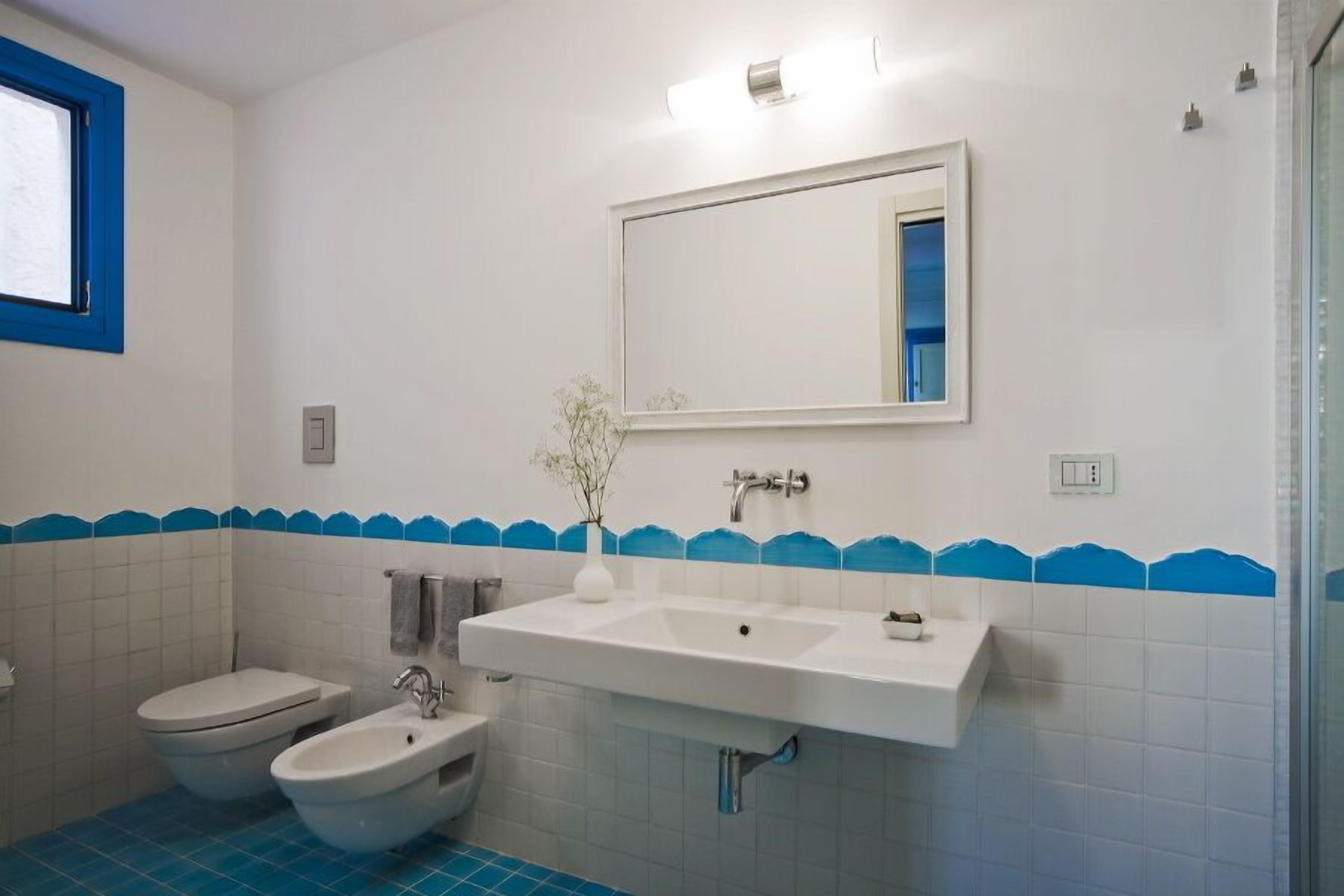 A bathroom with a white sink, two bidets, a toilet, and a mirror with a blue-tiled border.