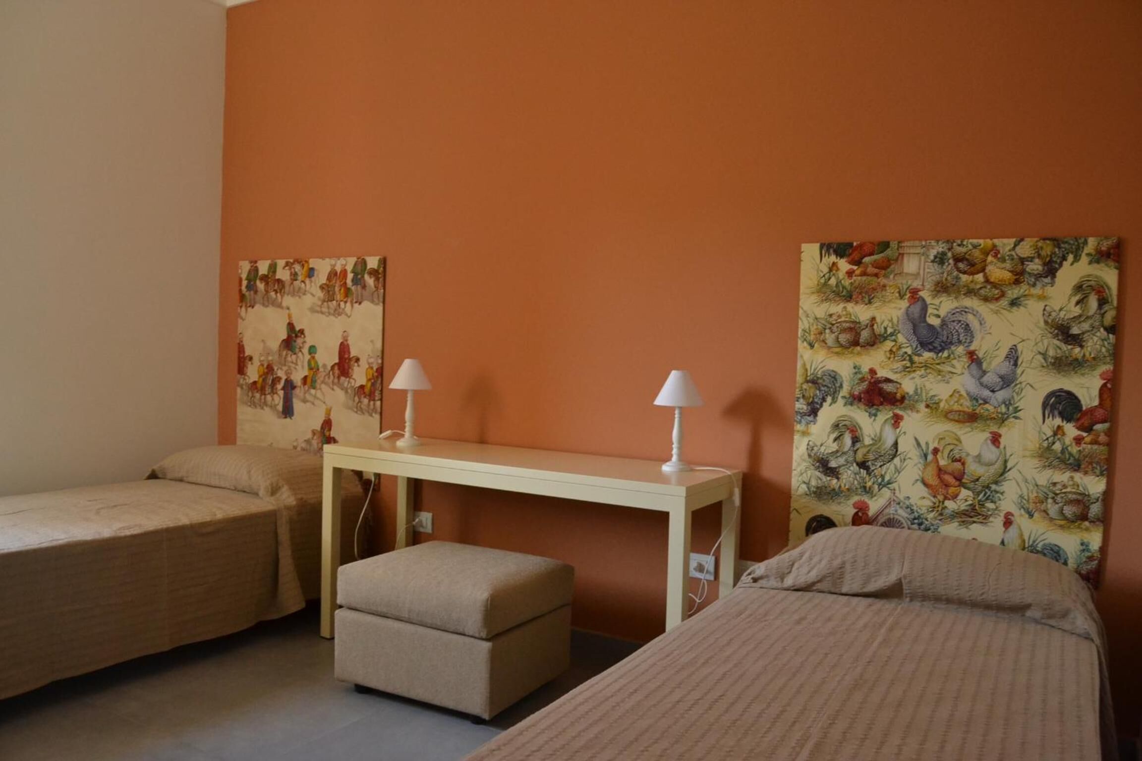 A room with two single beds, a small table with two lamps, and a wall with a rooster pattern.