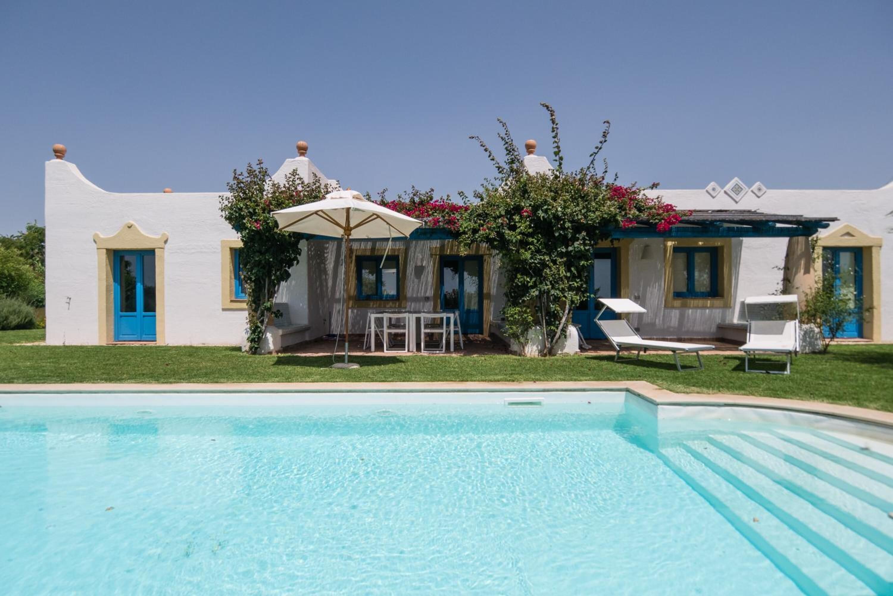 A poolside area with a white building, blue doors, and a table with umbrellas.