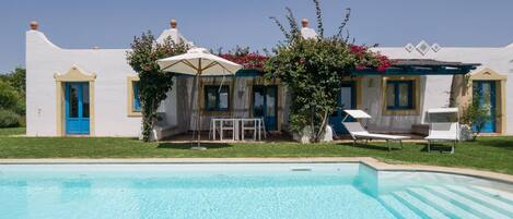 A poolside area with a white building, blue doors, and a table with umbrellas.
