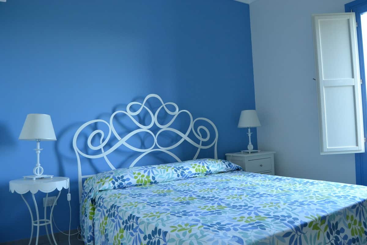 A bedroom with a blue wall, a white metal bed frame, a floral bedspread, two bedside tables with lamps, and a window with a white blind.