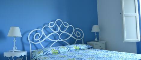 A bedroom with a blue wall, a white metal bed frame, a floral bedspread, two bedside tables with lamps, and a window with a white blind.