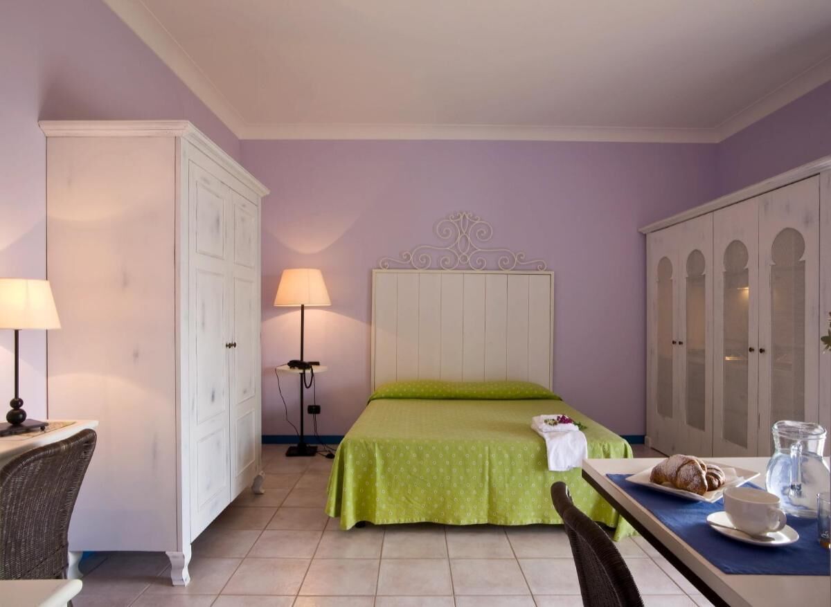 Quarto de hotel com cama, escrivaninha e cadeira, armário e mesa de cabeceira com abajur.
