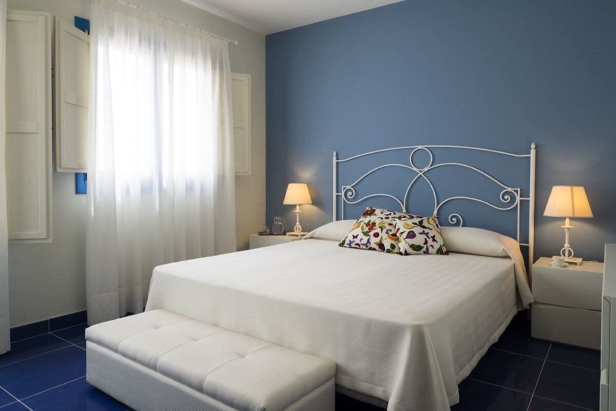 A bedroom with a bed, two bedside tables with lamps, a window with curtains, and a blue accent wall.