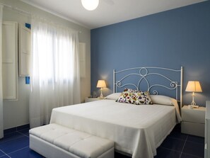 A bedroom with a bed, two bedside tables with lamps, a window with curtains, and a blue accent wall. - Marsa Siclà Le Ville (Scicli)