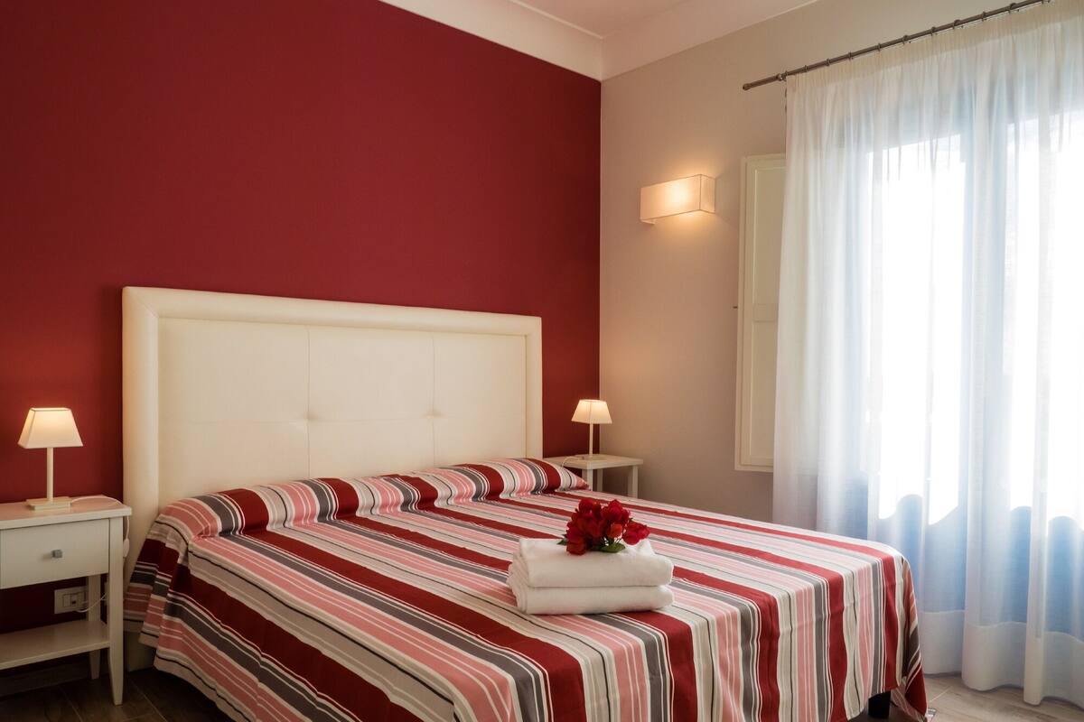 A hotel room with a large bed, two bedside tables, a nightstand, a window with sheer curtains, and a wall-mounted light.