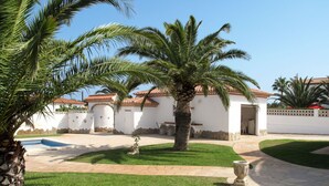 A white building with a tiled roof, a swimming pool, and a well-maintained lawn with palm trees.