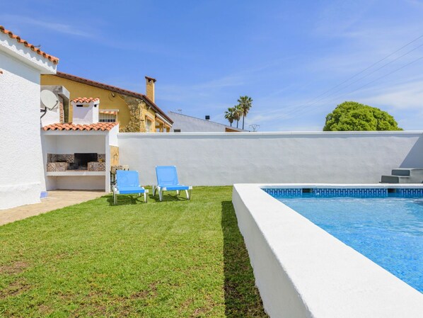 Una piscina con pared blanca y azulejos azules, dos sillas de descanso azules, y una zona ajardinada con un edificio al fondo.