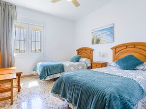 Un dormitorio con dos camas, una mesita de noche de madera, una mesa y una ventana con barrotes.