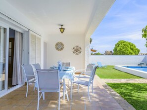 Un patio con una mesa dispuesta para cuatro personas, rodeado de sillas blancas, un muro decorativo y vistas a una piscina y follaje.