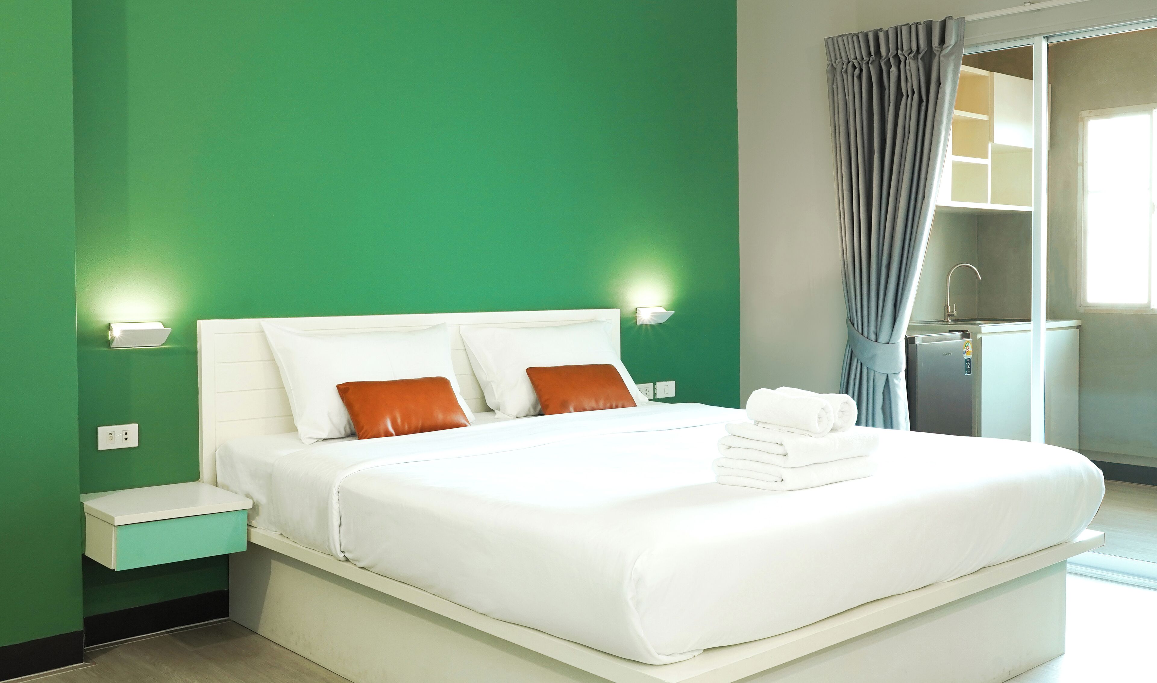 Een netjes opgemaakt bed met witte lakens en twee oranje kussens, een nachtkastje met een lamp en een groene muur.