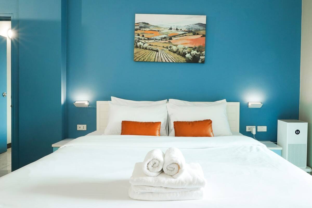 Ein Hotelzimmer mit einem großen Bett, weißer Bettwäsche, zwei orangefarbenen Kissen, einem Landschaftsbild an der Wand und einem gefalteten Handtuch auf dem Bett.
