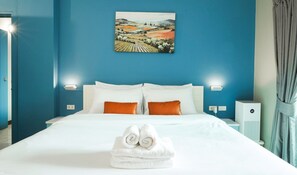 Ein Hotelzimmer mit einem großen Bett, weißer Bettwäsche, zwei orangefarbenen Kissen, einem Landschaftsbild an der Wand und einem gefalteten Handtuch auf dem Bett.