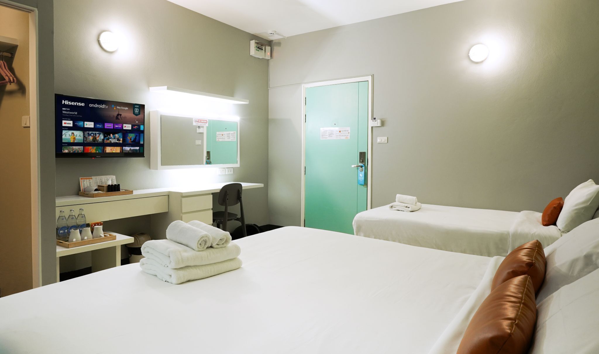 A hotel room with two beds, a desk with a computer, a TV displaying channels, and a door with a sign.