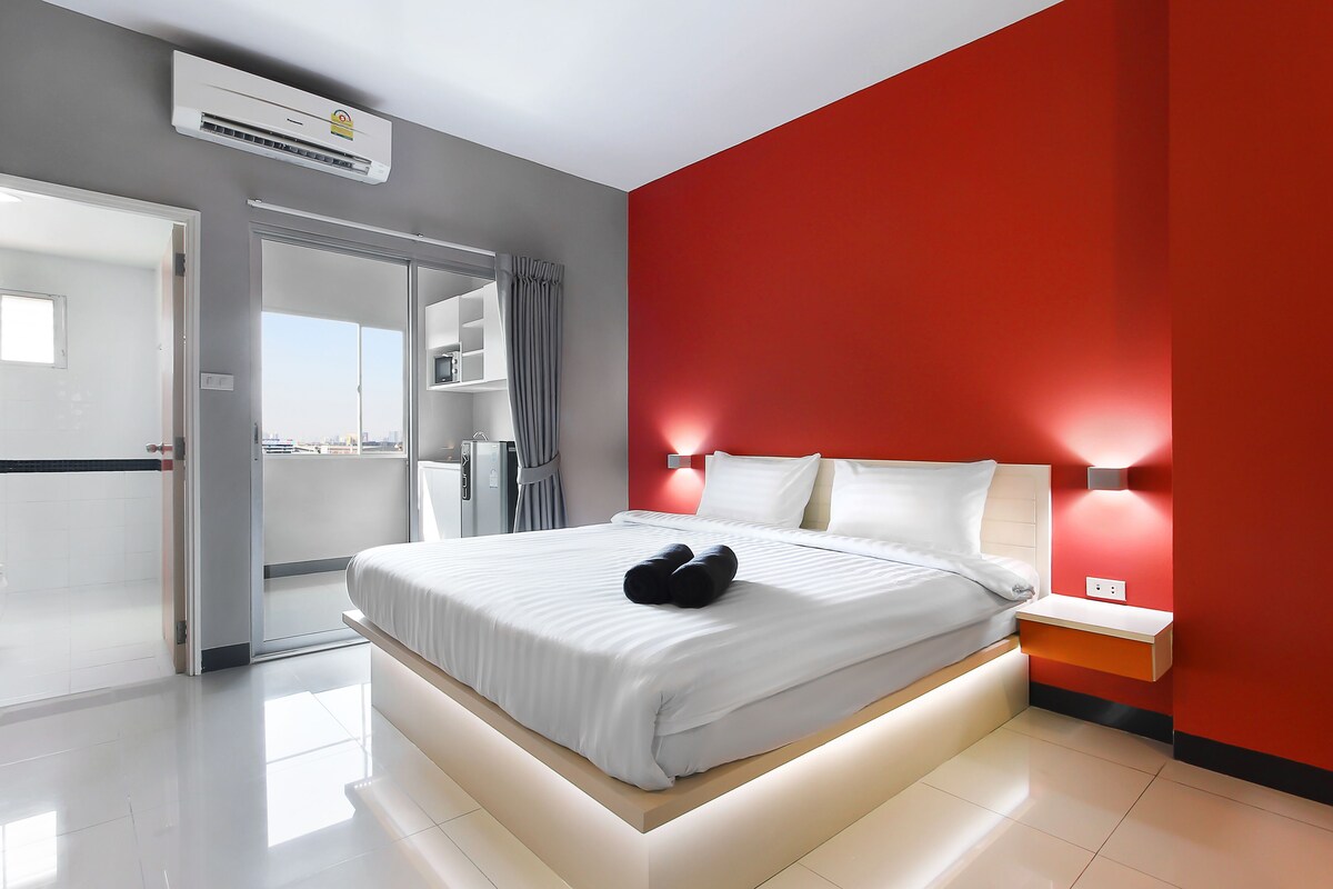 Une chambre d’hôtel moderne avec un grand lit, un mur d’accent rouge et une vue sur la ville, visible à travers la porte.