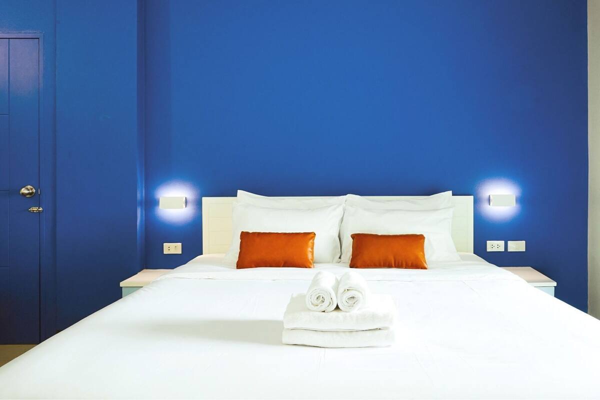 Ein Hotelzimmer mit einem großen Bett, weißen Leinentüchern, zwei orangefarbenen Kissen und einem ordentlich gefalteten Handtuch auf dem Bett.